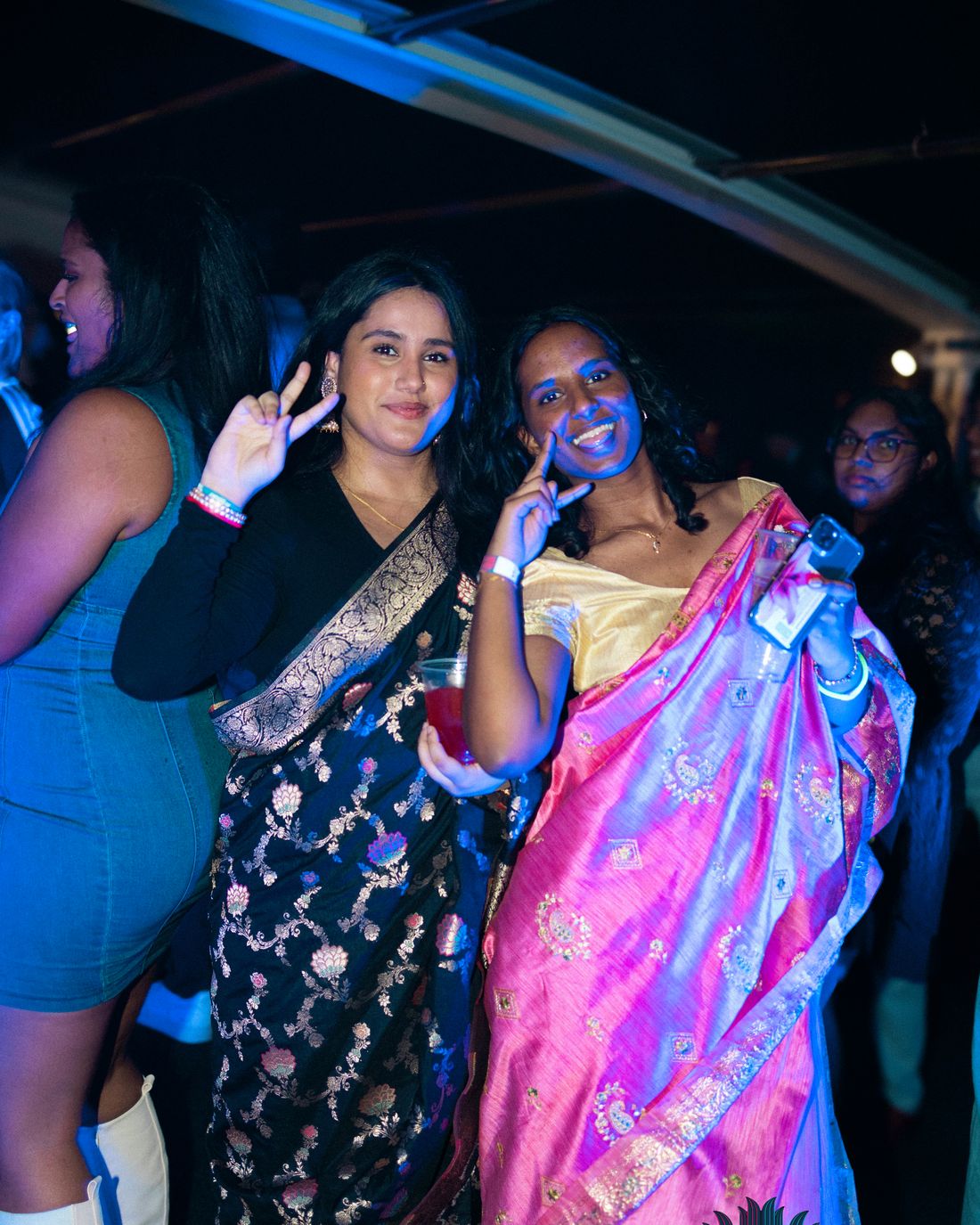 Two guests in sarees on the 2025 cruise, smiling at the camera under blue light, drink in hand.